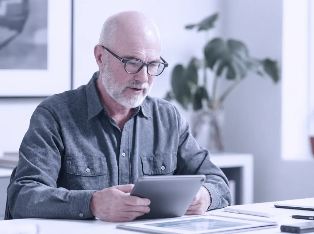 Älterer Mann mit Brille und grauem Hemd liest konzentriert auf einem Tablet in einem hellen, modernen Büro.